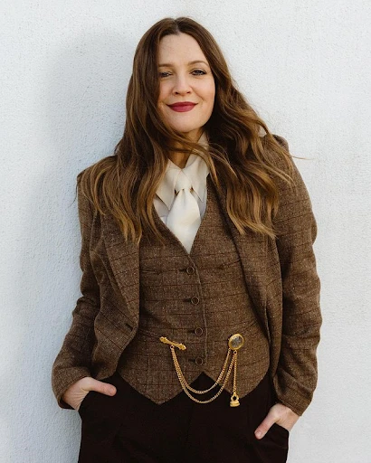Drew Barrymore wearing a brown tweed blazer, vest, and dark pants, posing against a white wall in a medium studio shot.