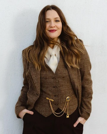 Drew Barrymore wearing a brown tweed blazer, vest, and dark pants, posing against a white wall in a medium studio shot.