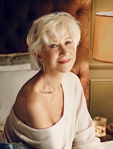 Close-up portrait of a smiling, mature woman with short white hair, wearing a cream sweater in a warm, inviting bedroom setting.