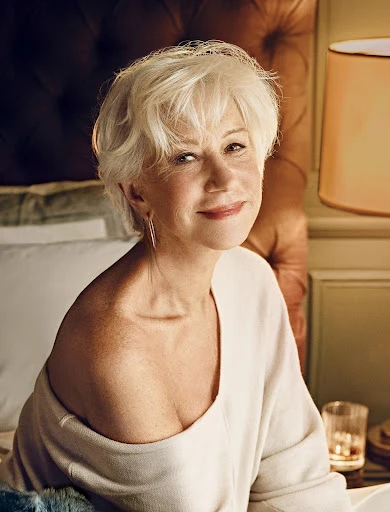 Close-up portrait of a smiling, mature woman with short white hair, wearing a cream sweater in a warm, inviting bedroom setting.