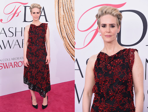 Sarah Paulson in a black and red floral dress on a pink carpet in front of a logoed backdrop at a fashion awards event.