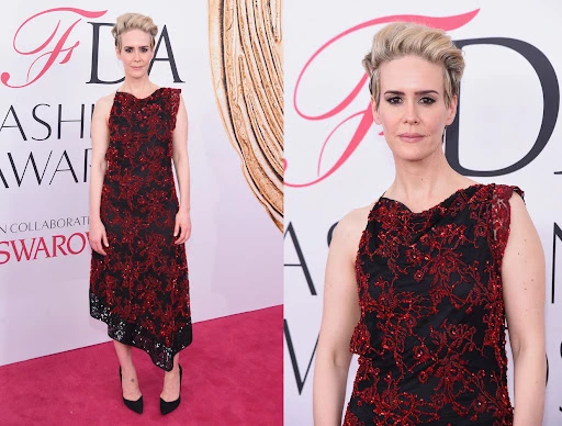 Sarah Paulson in a black and red floral dress on a pink carpet in front of a logoed backdrop at a fashion awards event.