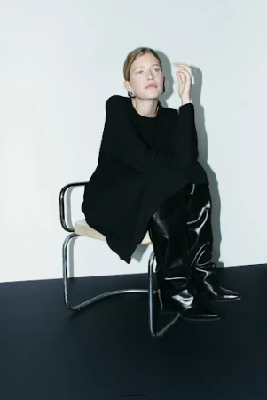 Adult female model poses in a photo studio, wearing a black top, skirt, and boots, with a red background.