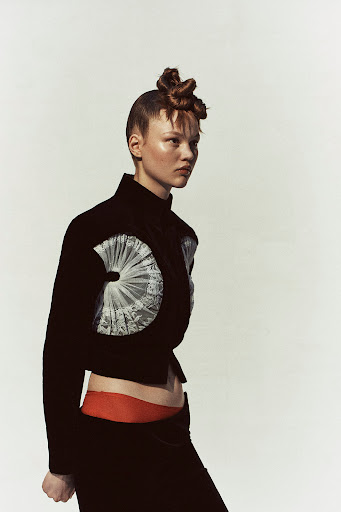 A stylish young woman poses in a black jacket with an elaborate lace detail, set against a clean, light background for a high-fashion editorial shot.