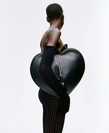 A model with dark skin stands sideways against a white background, wearing a textured dress and gloves, and carrying a heart-shaped black bag.