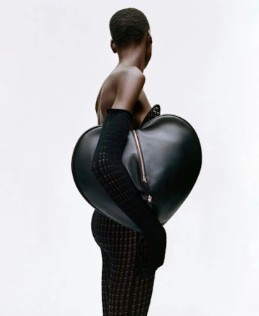A model with dark skin stands sideways against a white background, wearing a textured dress and gloves, and carrying a heart-shaped black bag.