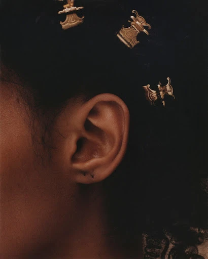 Close-up of an adult female's ear with ornate, gold-toned jewelry in dark curly hair against a dark background in a studio setting.