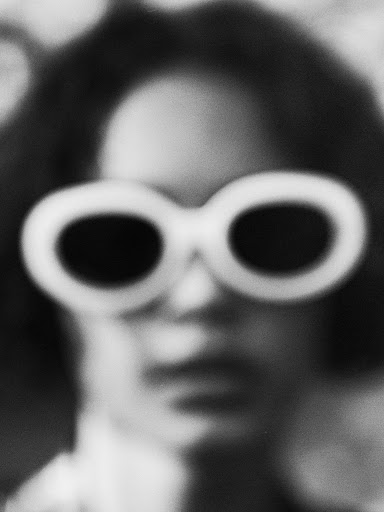 Close-up portrait of a person wearing large white-rimmed sunglasses in a grainy monochrome aesthetic.
