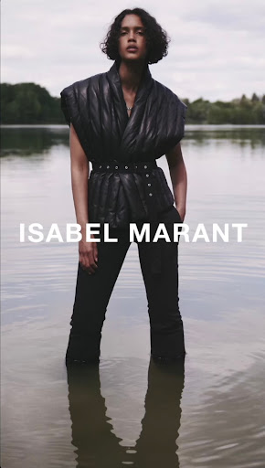 An adult fashion model poses in water wearing a black quilted vest, matching trousers, and a belt. The sky is overcast.