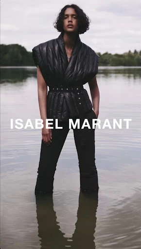 An adult fashion model poses in water wearing a black quilted vest, matching trousers, and a belt. The sky is overcast.