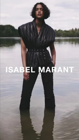 An adult fashion model poses in water wearing a black quilted vest, matching trousers, and a belt. The sky is overcast.