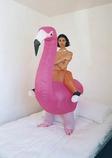 An adult female poses in a pink flamingo costume on a white bed in front of a white wall with soft lighting and a portrait orientation.
