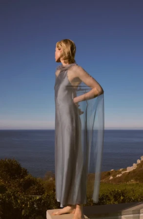 A lone woman in a flowing grey gown poses against a sea and sky backdrop, showcasing a blend of natural beauty and high fashion.