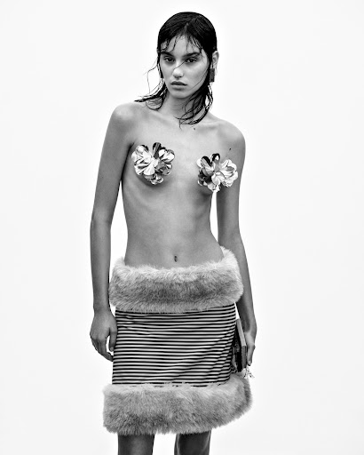 Black and white studio shot of a slender young woman wearing a striped mini-skirt with fur trim, and flowers covering her chest.