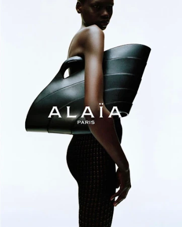 Black model holding a large structured black leather handbag against a plain white background with brand text ALAIA PARIS