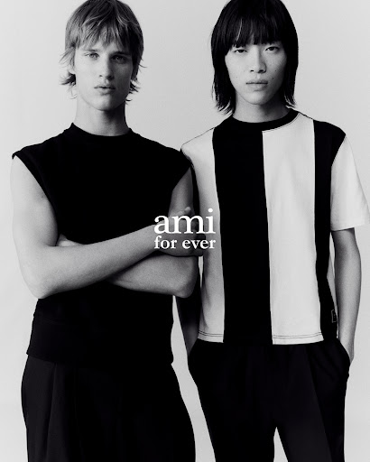 Two adult male fashion models stand together indoors wearing black and white t-shirts with a grey background in a studio shot.
