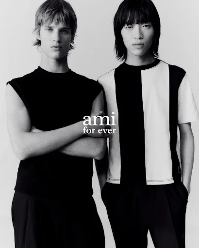 Two adult male fashion models stand together indoors wearing black and white t-shirts with a grey background in a studio shot.