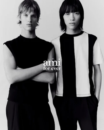 Two adult male fashion models stand together indoors wearing black and white t-shirts with a grey background in a studio shot.