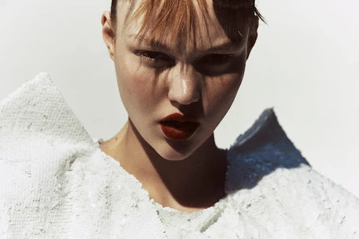 Close-up portrait of a woman with light skin, dark red lipstick, and a sequined garment against a bright, neutral background.