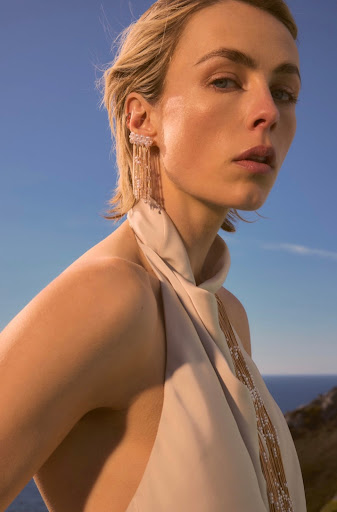 Blond woman with halter dress and statement earrings posing outdoors against a blue sky and coastal scenery in a medium shot.