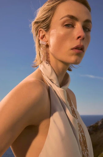 Blond woman with halter dress and statement earrings posing outdoors against a blue sky and coastal scenery in a medium shot.