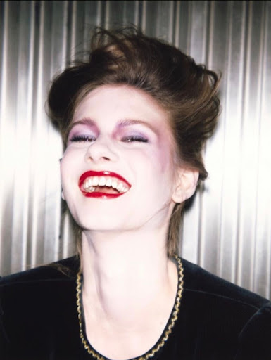 Headshot of a happy woman with a black top and bright red lipstick in front of a gray corrugated metal background.