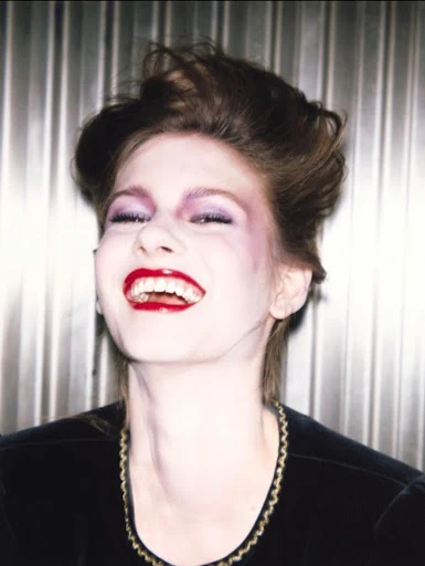 Headshot of a happy woman with a black top and bright red lipstick in front of a gray corrugated metal background.