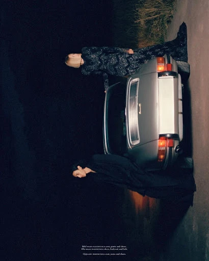 Two models pose with a silver car on a dark road at night, wearing elegant dresses in a fashion editorial shot with low-key lighting.