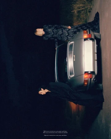 Two models pose with a silver car on a dark road at night, wearing elegant dresses in a fashion editorial shot with low-key lighting.