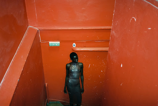 Fashion photograph of a black female model wearing a fitted dress in a red staircase with a bright exit sign above her head.