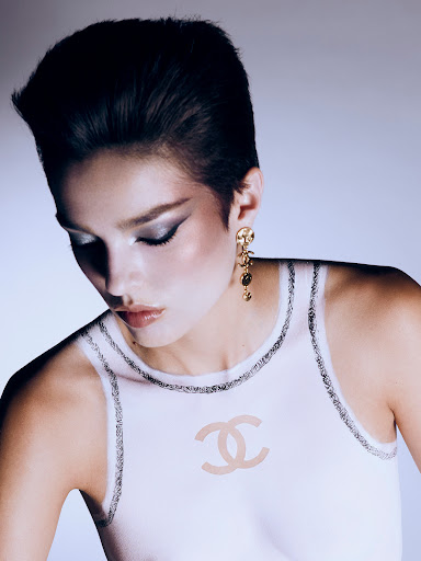 Close-up of a fashionable young adult woman with short dark hair and grey eyeshadow wearing a white top with the Chanel logo.