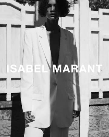 Monochrome portrait of a model with short curly hair wearing an oversized white blazer against a patterned fence.