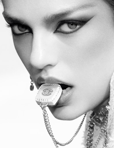Close up portrait of a woman wearing dramatic winged eyeliner holding a branded logo charm with a chain in her teeth