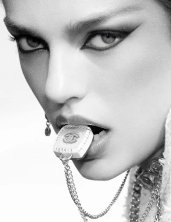 Close up portrait of a woman wearing dramatic winged eyeliner holding a branded logo charm with a chain in her teeth