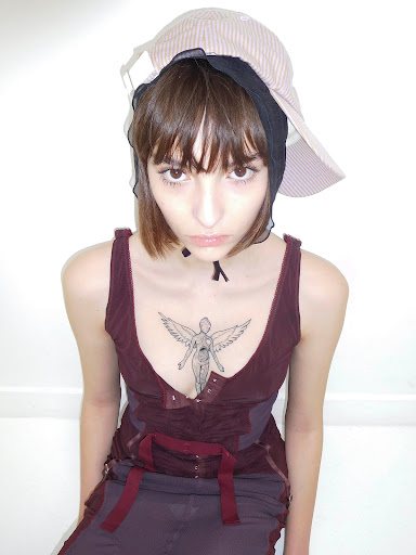 Young woman with short brown hair wears a maroon top and layered hats in a brightly lit indoor portrait, showcasing alternative fashion.