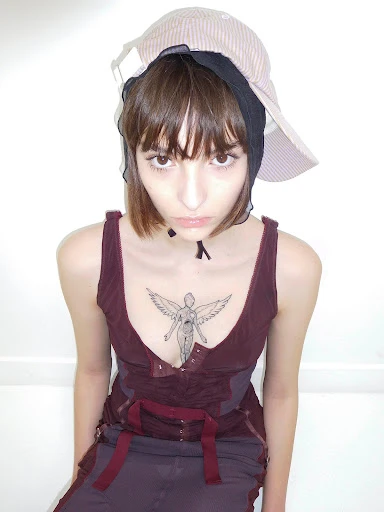 Young woman with short brown hair wears a maroon top and layered hats in a brightly lit indoor portrait, showcasing alternative fashion.