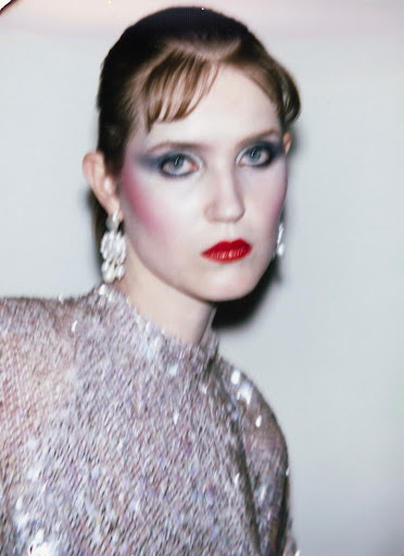 Portrait of an adult female with red lips and heavy blue eyeshadow, wearing a sequined top and chandelier earrings against a neutral background.