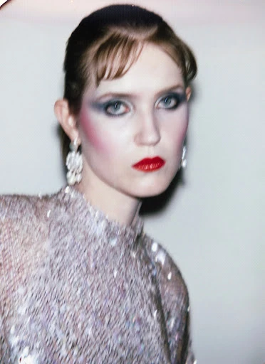 Portrait of an adult female with red lips and heavy blue eyeshadow, wearing a sequined top and chandelier earrings against a neutral background.