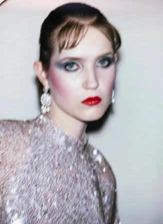 Portrait of an adult female with red lips and heavy blue eyeshadow, wearing a sequined top and chandelier earrings against a neutral background.