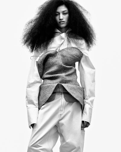 Fashion shot of a woman with large hair, wearing a vest over a white shirt, and white pants against a bright background.