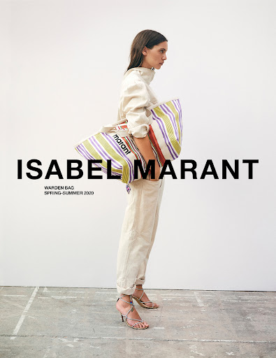 Fashion editorial shot of a female model carrying a striped tote bag with the brand logo, posing against a neutral background indoors.