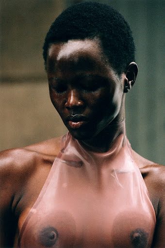 Close-up of a topless Black woman with a lip ring and short hair, wearing a translucent pink halter top, posing in front of a concrete wall.