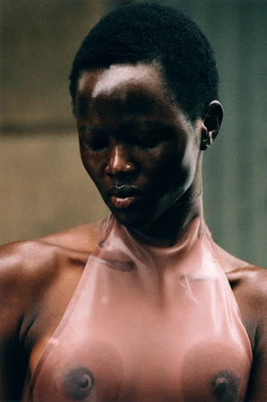 Close-up of a topless Black woman with a lip ring and short hair, wearing a translucent pink halter top, posing in front of a concrete wall.