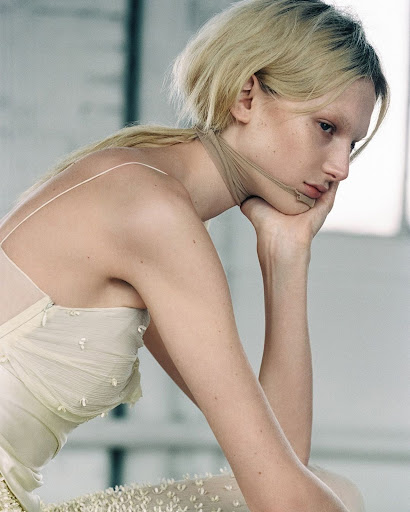 Pale blonde fashion model poses in a studio setting, showcasing a sheer dress with textured floral accents and a delicate tulle scarf.