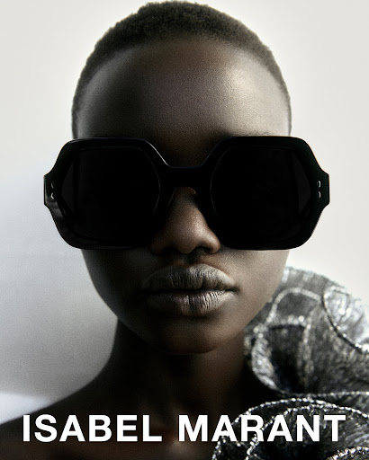 Portrait of a dark-skinned fashion model wearing black sunglasses and a shiny silver dress with the words Isabel Marant on the bottom.