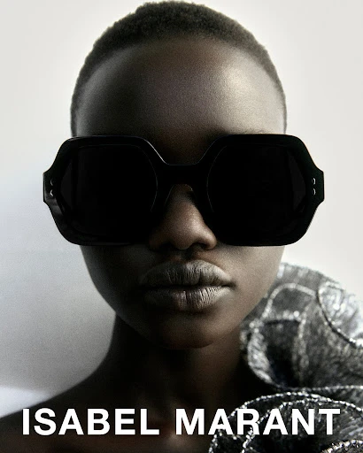 Portrait of a dark-skinned fashion model wearing black sunglasses and a shiny silver dress with the words Isabel Marant on the bottom.