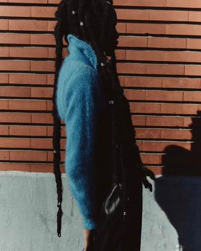 Stylish woman with dreadlocks and a blue sweater posing against a brick wall in natural light, showcasing fashion and texture.