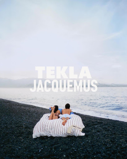 A couple lies on a striped mattress on a black pebble beach next to the ocean under a cloudy sky with the words TEKLA JACQUEMUS.