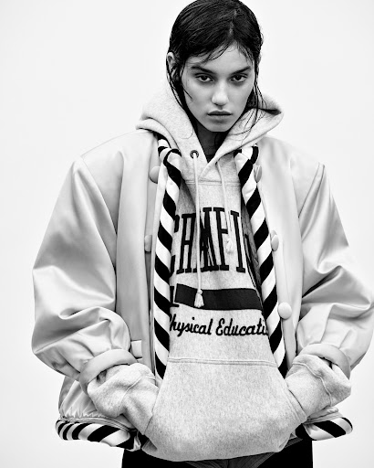 Monochrome portrait of a woman in a hoodie and jacket, with a white background, fashion-forward style, and soft lighting.