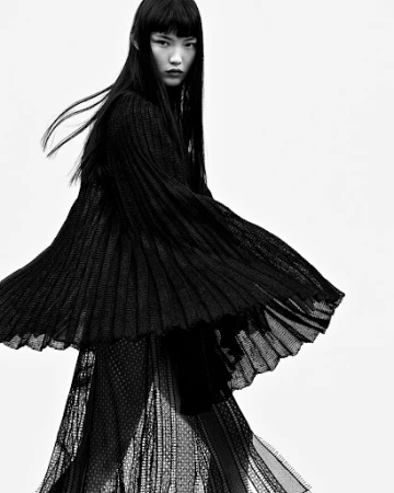 Model with long dark hair wearing a textured black pleated dress against a plain white background in Paris.